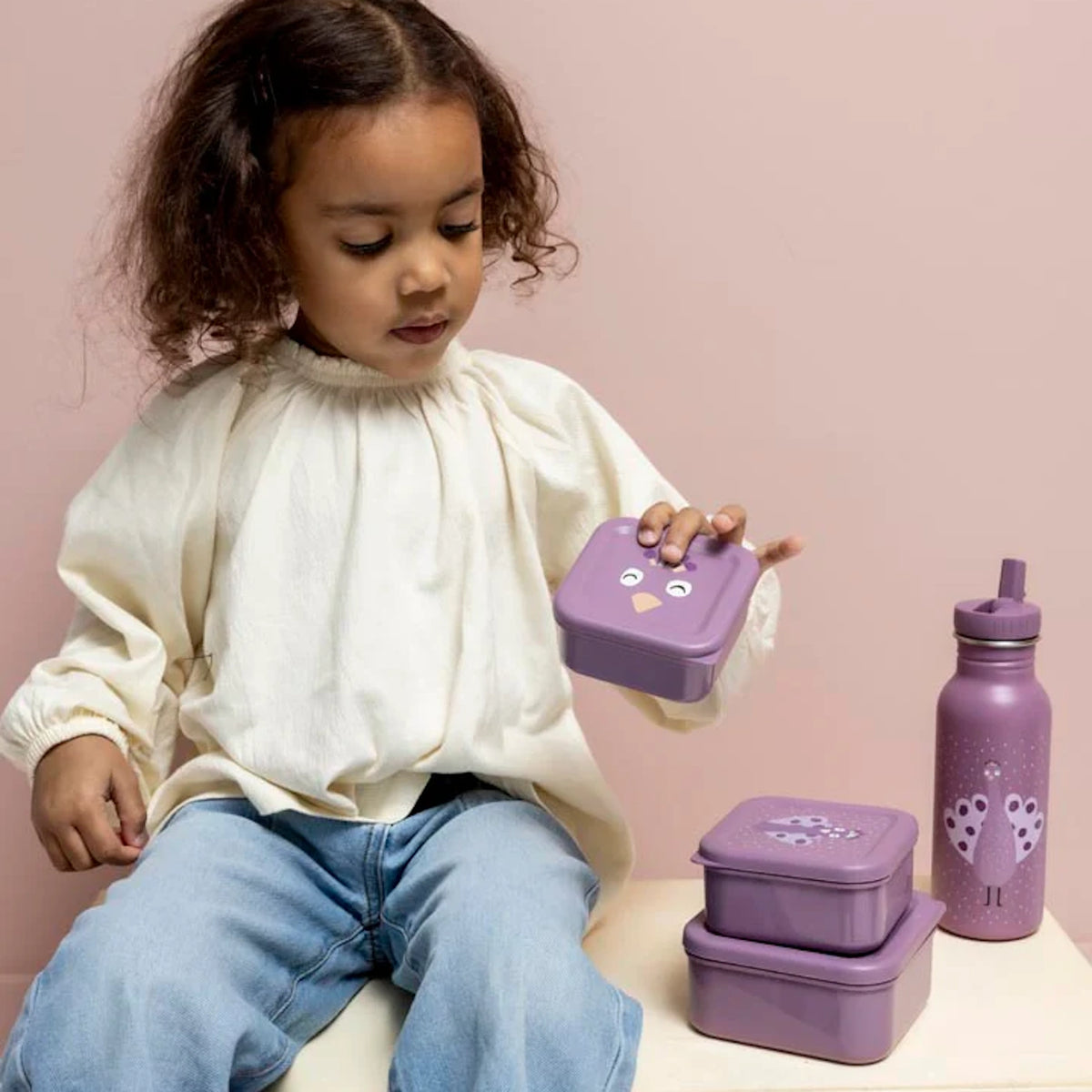 A young girl holding a purple peacock snack box next to matching nesting containers and a water bottle.