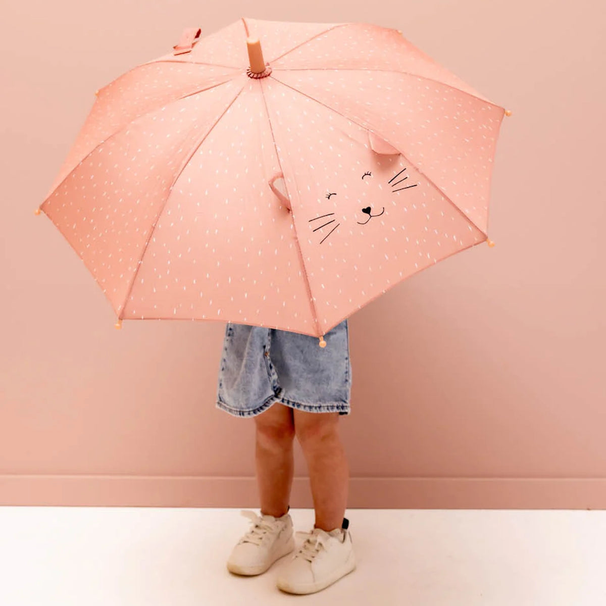A child holding a pink umbrella with a cartoon cat face, fabric ears, and white specks.