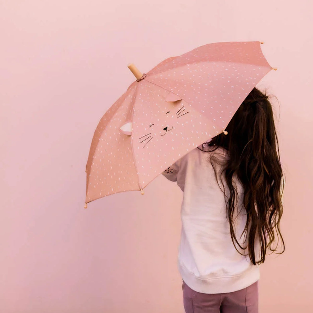 A child with long hair holding a pink umbrella with a cartoon cat face and pop-out ears against a pink wall.