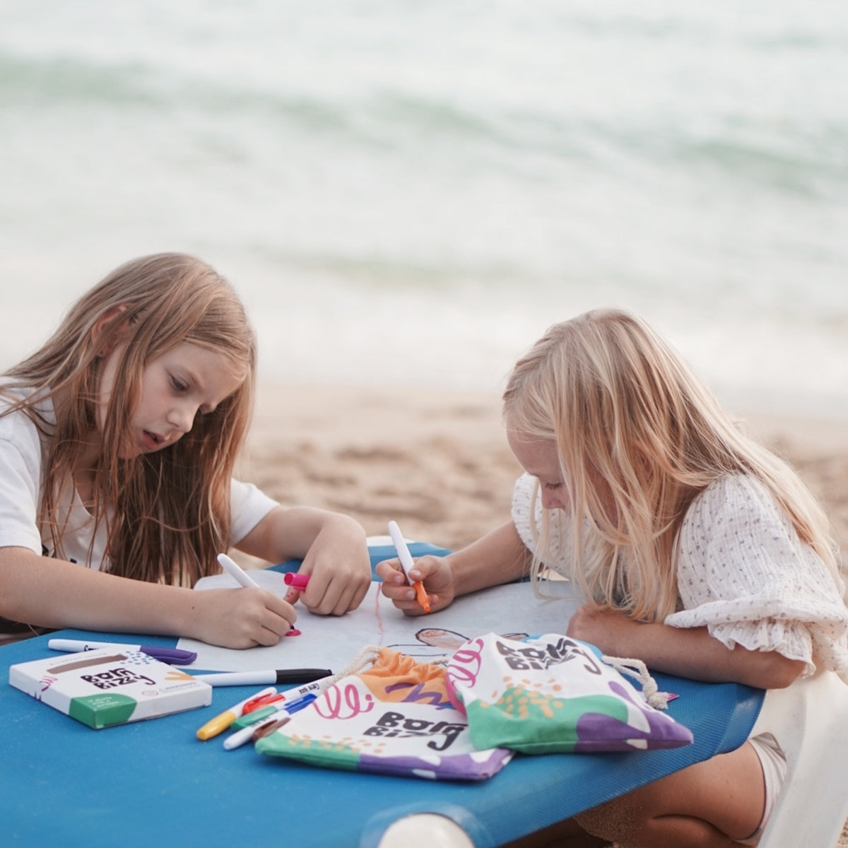 Two girls sit at a blue table on a sandy beach, focused on coloring with markers on a large sheet of paper.