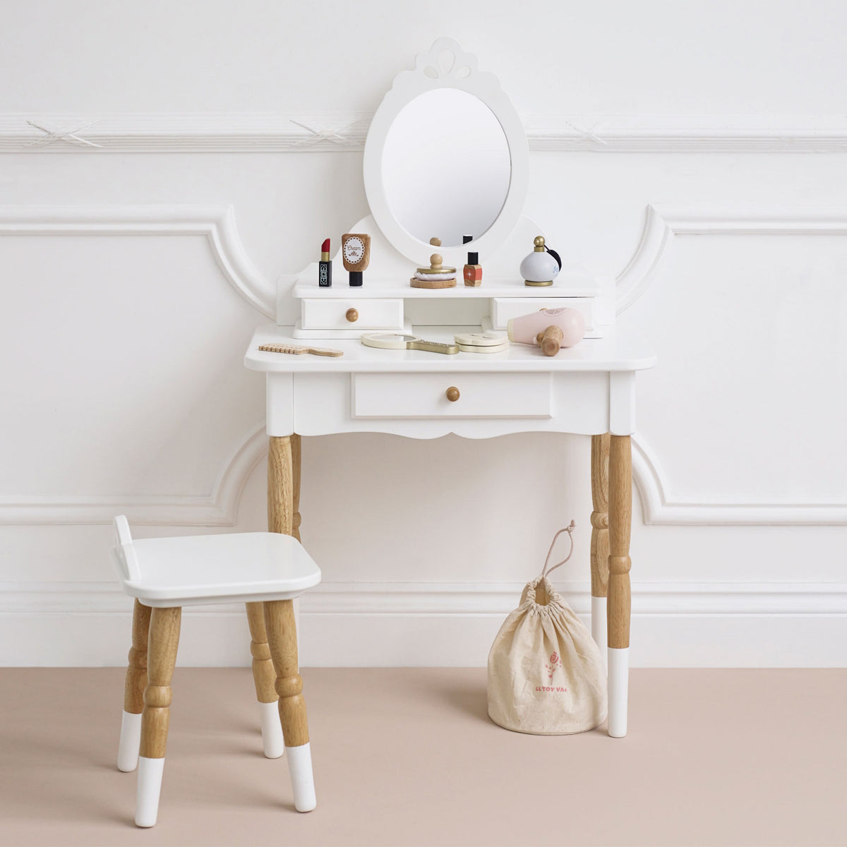 White wooden toy vanity with oval mirror, stool, play makeup accessories, and a storage bag.