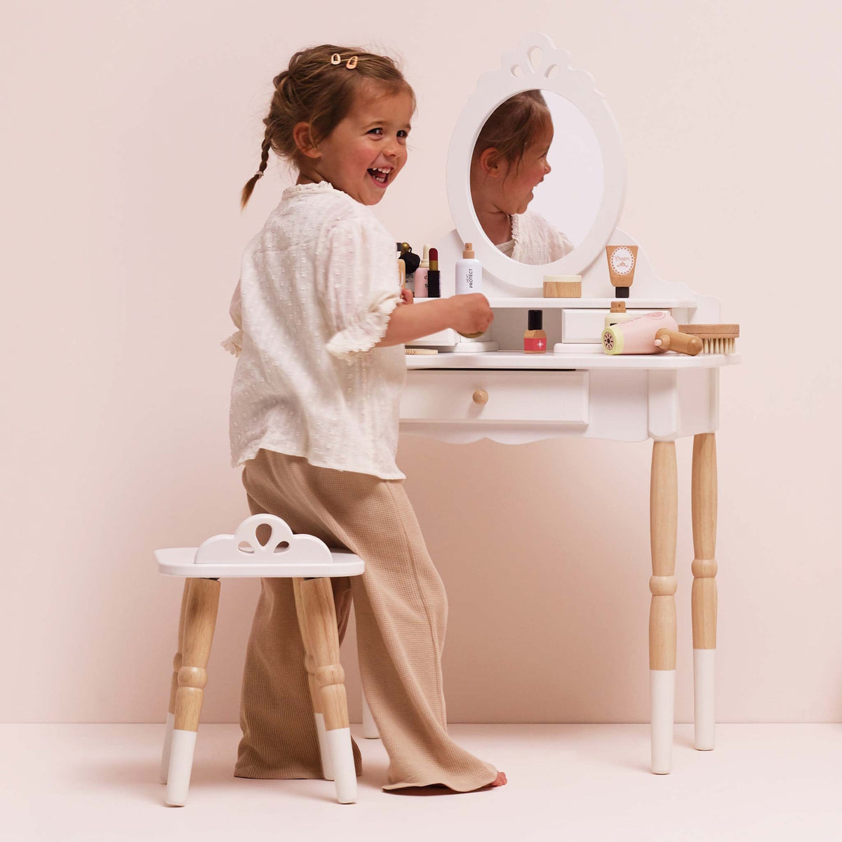 Smiling child playing with wooden toy cosmetics at a white vanity table set with a stool.
