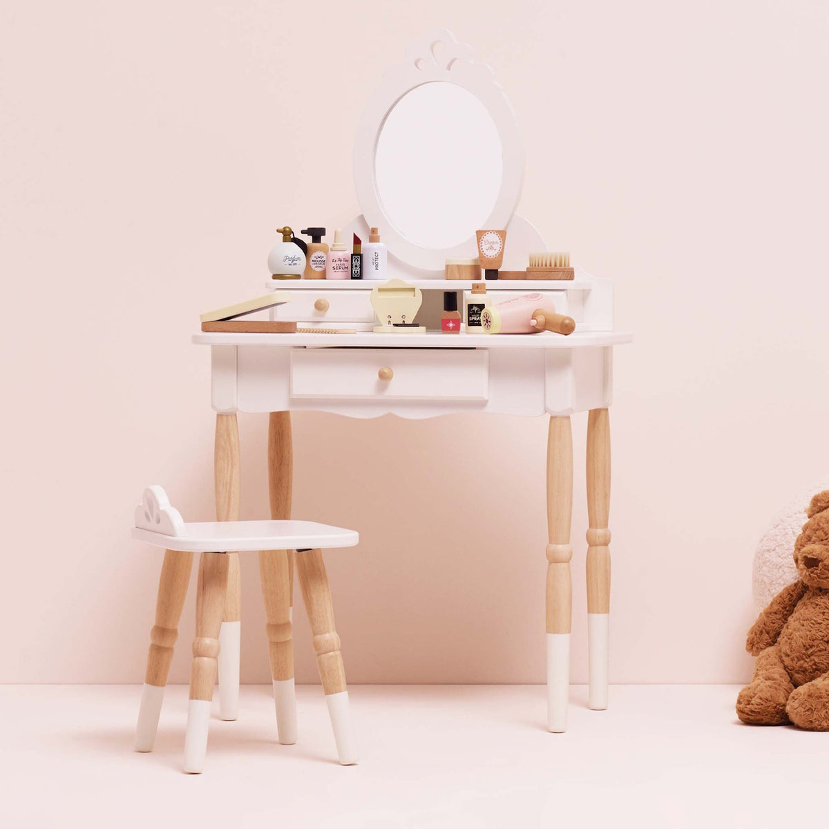 White wooden toy vanity table and stool set with play cosmetics, mirror, and a teddy bear nearby.