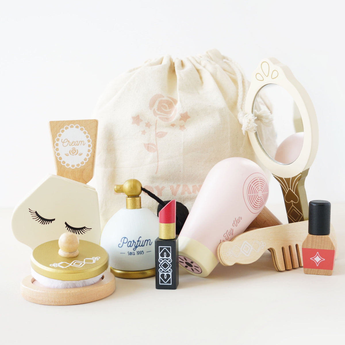 Wooden beauty play set with mirror, hair dryer, perfume, and makeup arranged in front of a cloth bag.