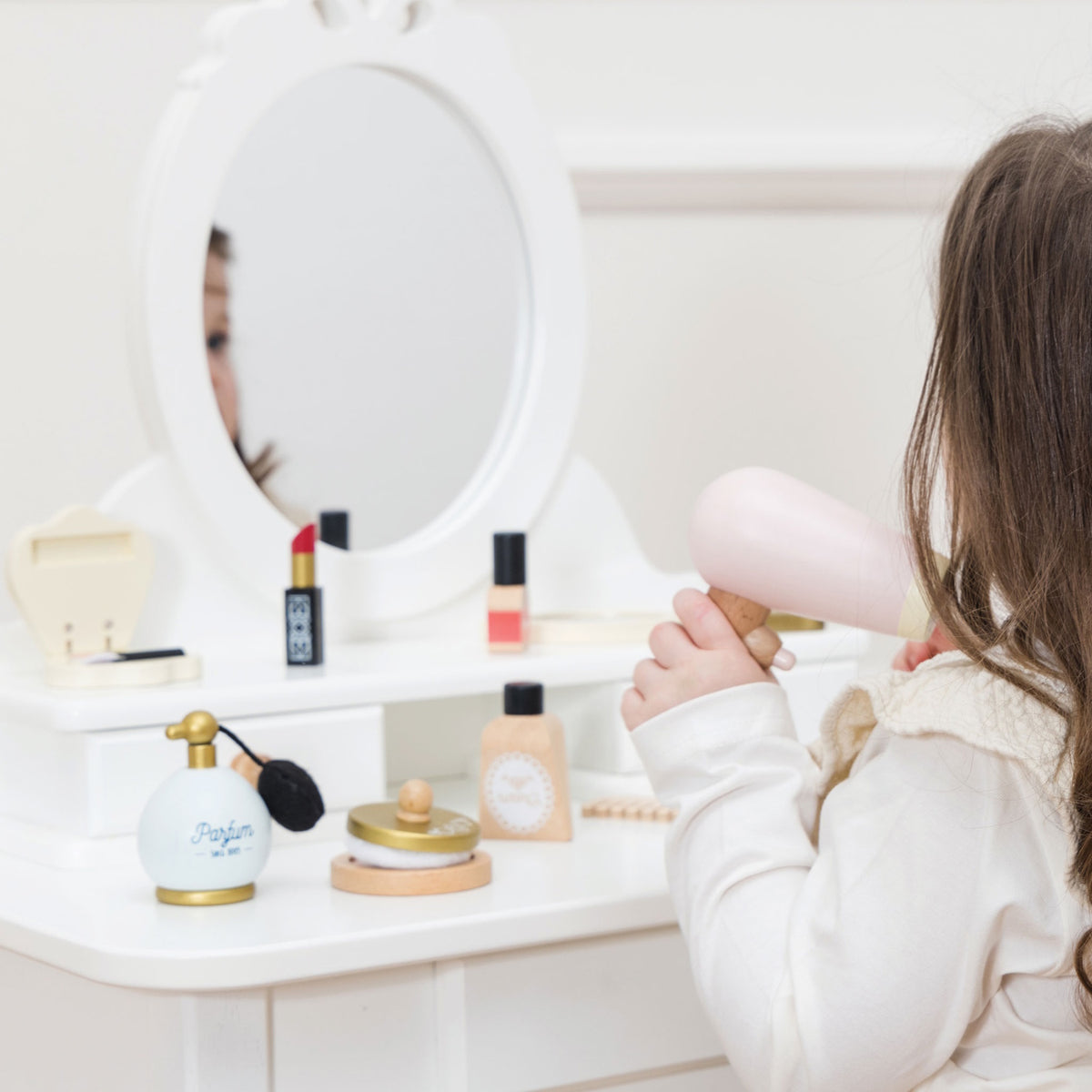 Child using a toy wooden hair dryer at a white vanity table set with play cosmetics.