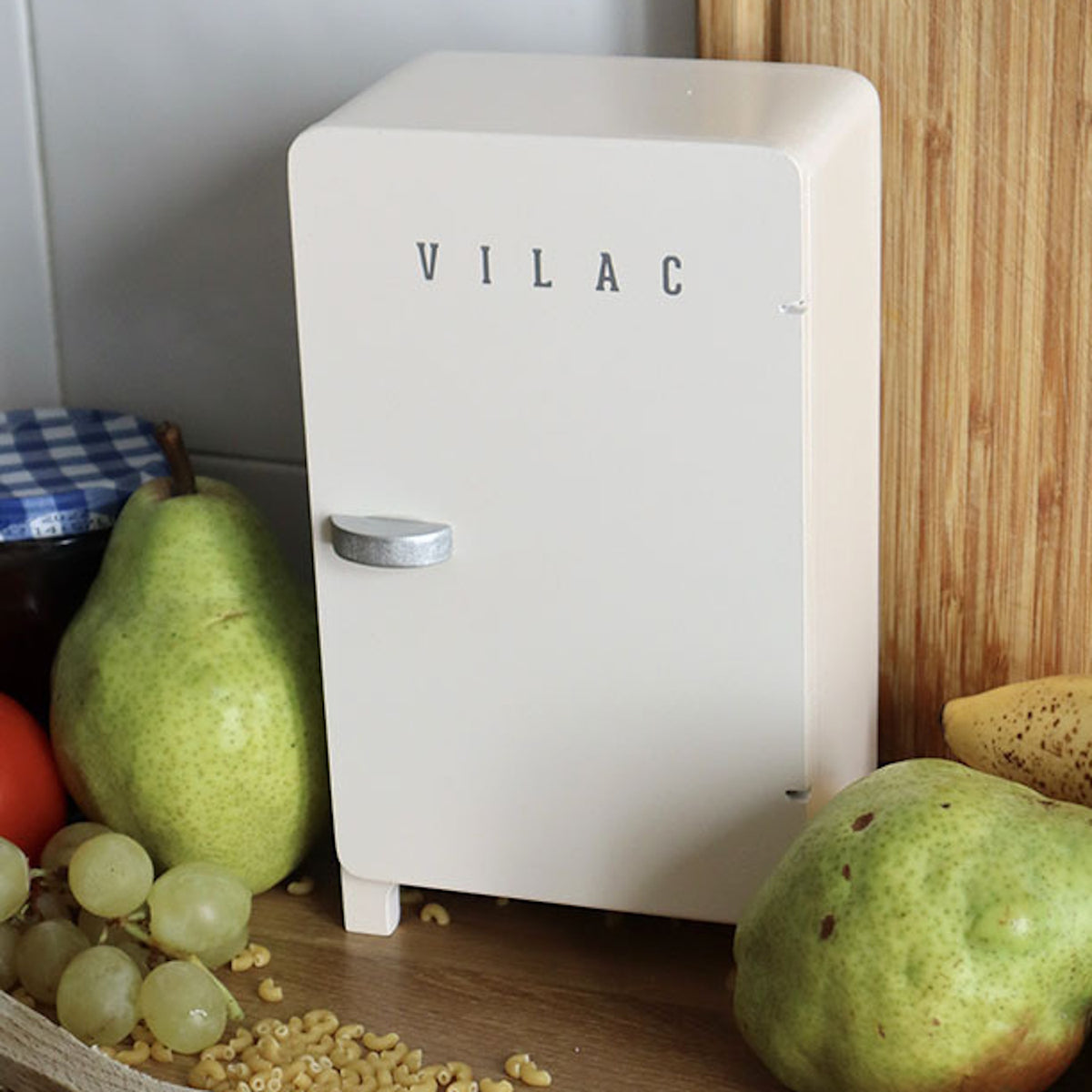 A cream Vilac toy fridge surrounded by fresh pears, grapes, and a banana on a wooden countertop.
