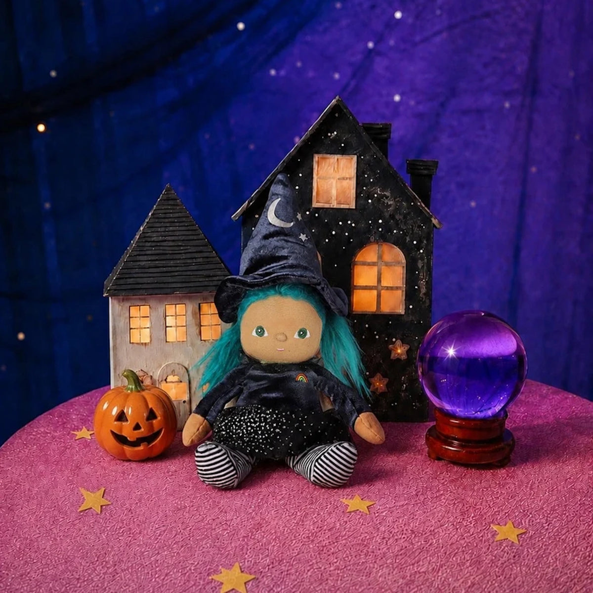 Plush witch doll sitting next to a glowing purple crystal ball, jack-o'-lantern, and miniature houses.