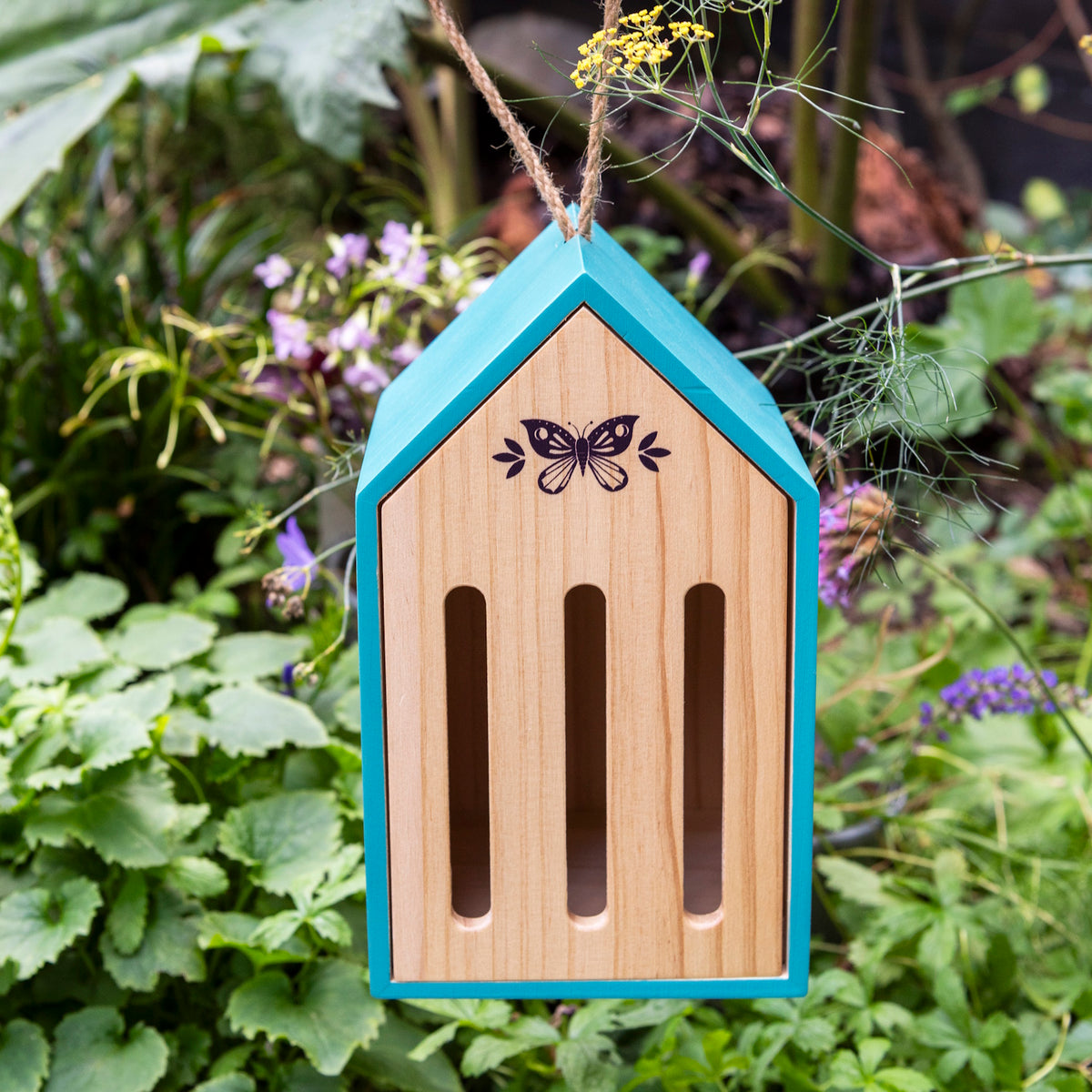 Teal wooden butterfly hotel hanging by a twine rope in a lush garden surrounded by green leaves and purple flowers.
