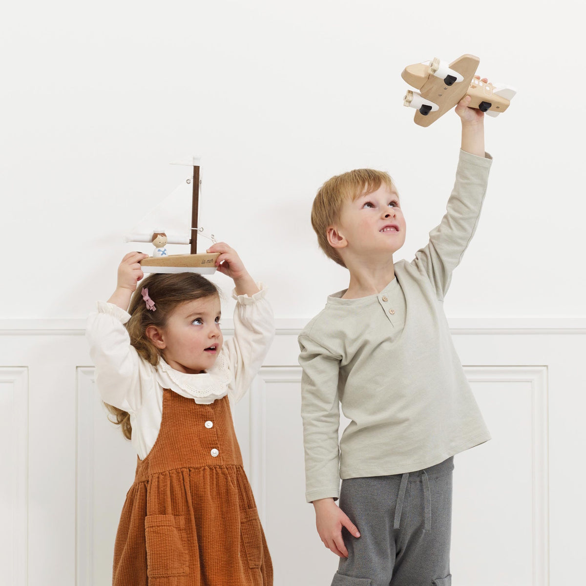 Young girl holding a wooden toy boat on her head next to a boy raising a wooden toy plane.