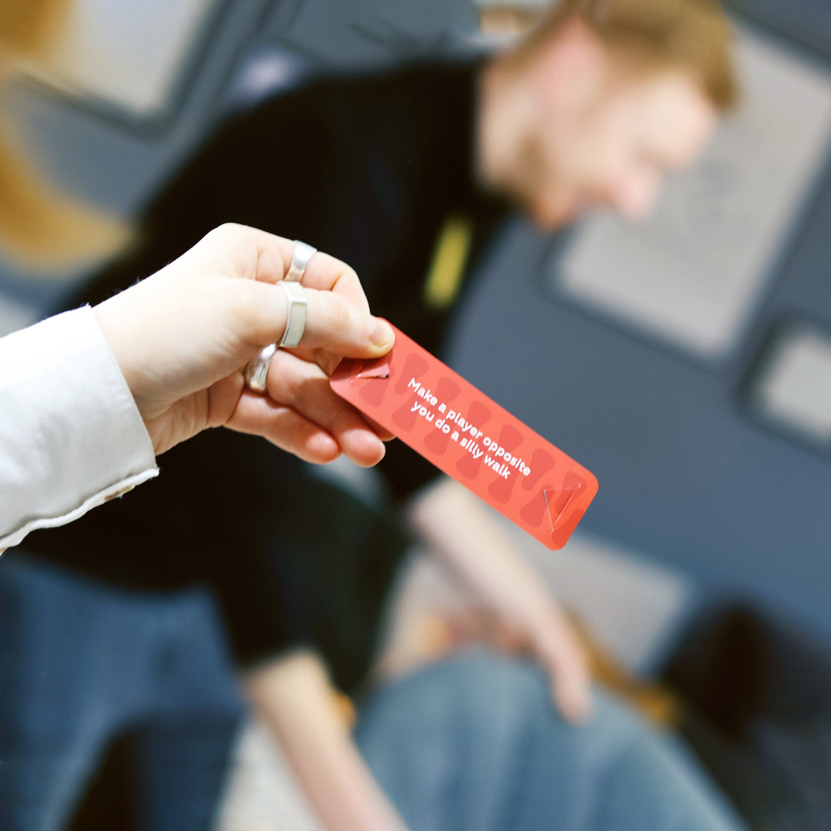A person holding a red Zip-it! game card with text reading "Make a player opposite you do a silly walk".