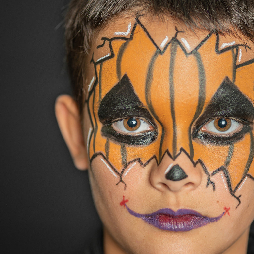 A close-up of a child with a Halloween-inspired pumpkin face paint design, featuring jagged black details and a purple lip colour.