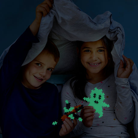 Two smiling children under a blanket holding glowing Plus-Plus creations, including a ghost and a rocket.