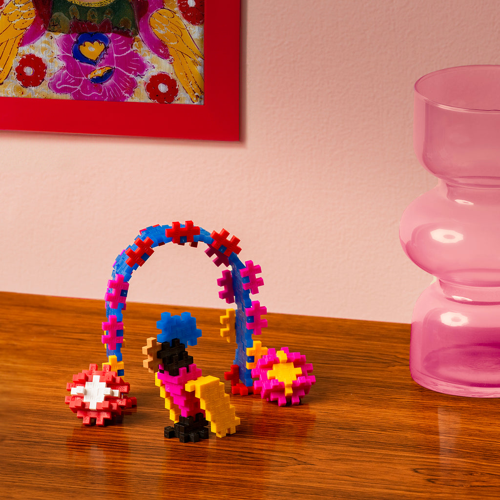 A colourful Plus-Plus construction inspired by Frida Kahlo's artwork, displayed on a wooden table with a pink glass vase in the background.