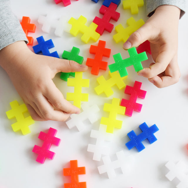 A close-up of a child's hands grasping colourful oversized Plus-Plus pieces in various neon and primary colours.