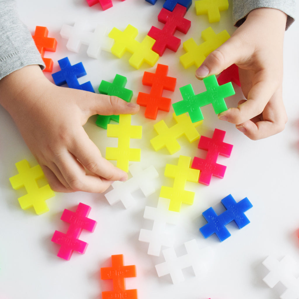 A close-up of a child's hands grasping colourful oversized Plus-Plus pieces in various neon and primary colours.