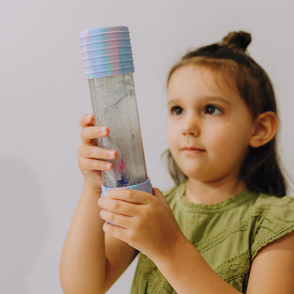 A young girl in a green dress holding and examining a Jellystone DIY Calm Down Bottle in pastel unicorn colours.