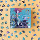 A whimsical Londji My Mermaid glitter puzzle box surrounded by scattered puzzle pieces on a yellow background.