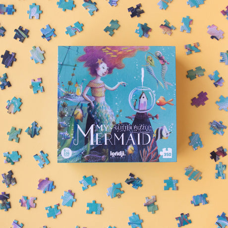 A whimsical Londji My Mermaid glitter puzzle box surrounded by scattered puzzle pieces on a yellow background.