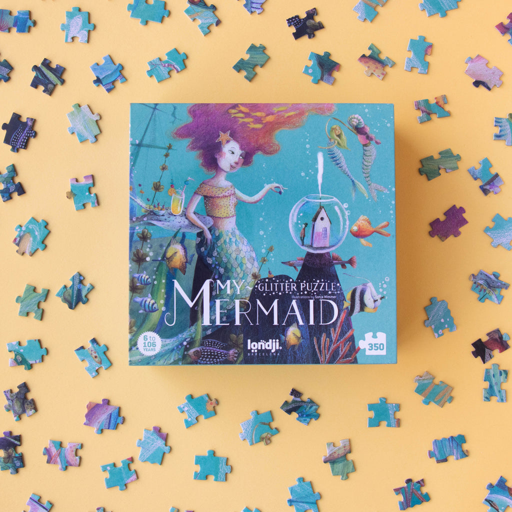 A whimsical Londji My Mermaid glitter puzzle box surrounded by scattered puzzle pieces on a yellow background.