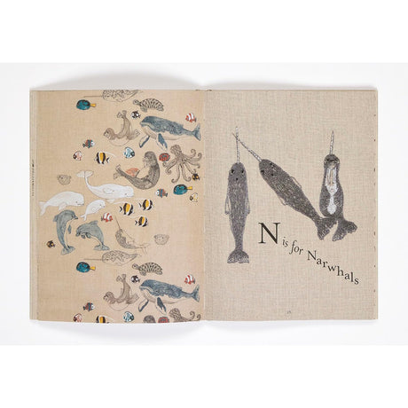 Children's book spread with sea creatures on the left and "N is for Narwhals" on linen-texture paper.