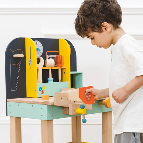 A young boy in a white shirt plays with a colorful wooden toy workbench, featuring tools and a saw. The scene conveys curiosity and engagement.