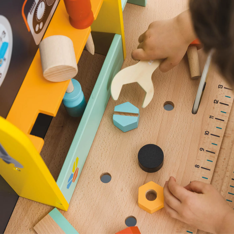 Child plays on a wooden toy workbench, using a wrench on a blue hexagon block. Tools, colorful shapes, and a ruler add a playful, creative feel.