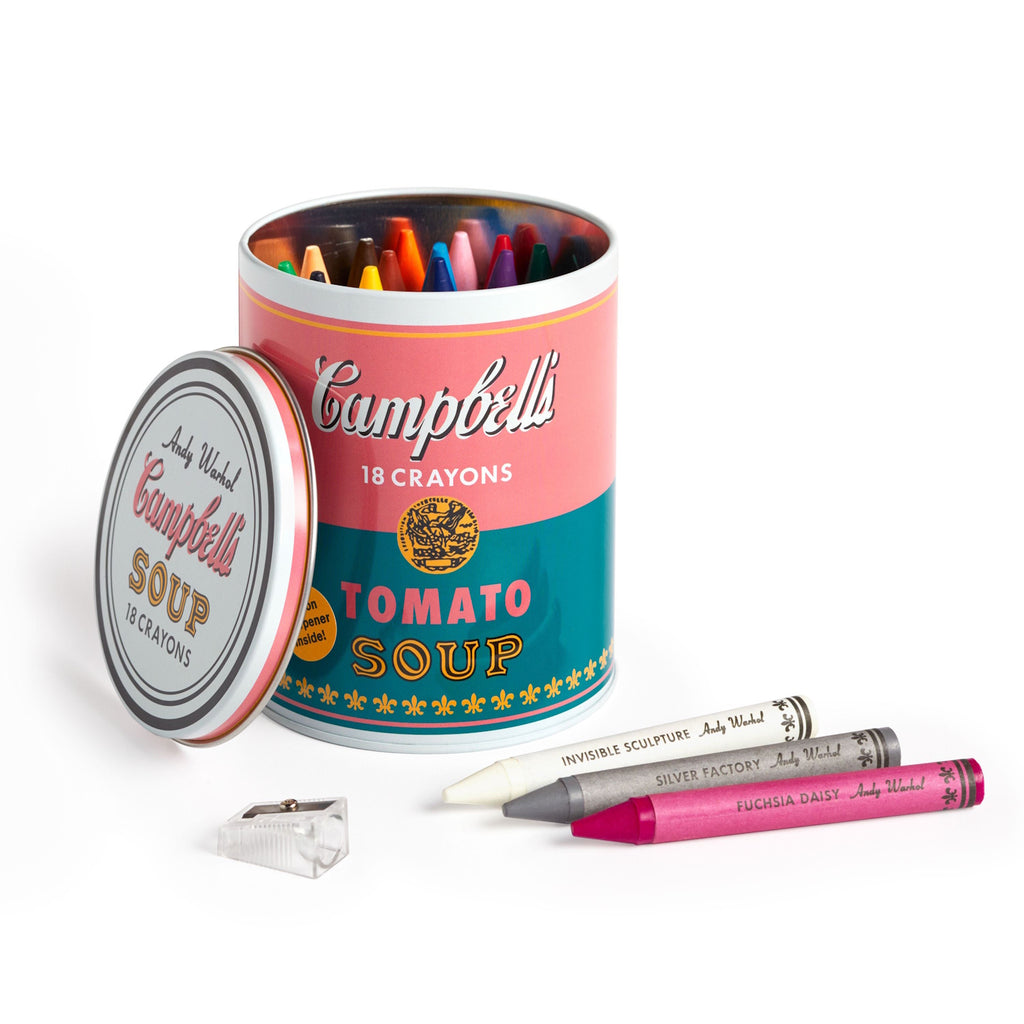Campbell's soup can with 18 colorful crayons, lid detached beside three crayons in white, gray, and pink. A small sharpener lies nearby. Playful, artistic tone.