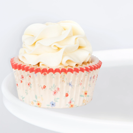 Frosted cupcake in a floral paper liner with a scalloped red rim on a white stand.