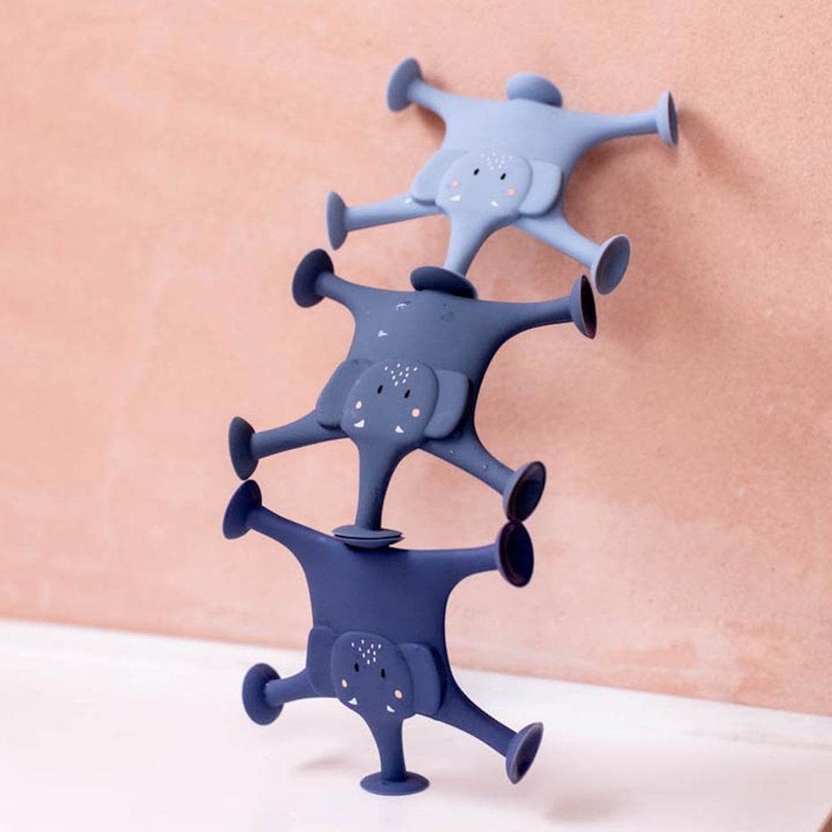 Three stackable elephant-shaped suction toys in shades of blue are attached to a peach wall. The setup is playful and imaginative.