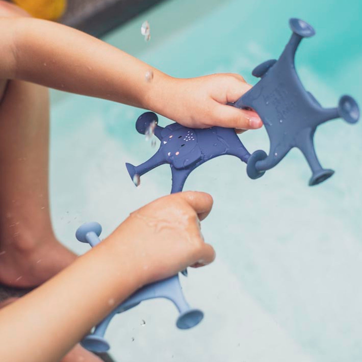 Child's hands hold blue toy starfish with suction cups above a pool. Water droplets splash, evoking a playful and cheerful atmosphere.