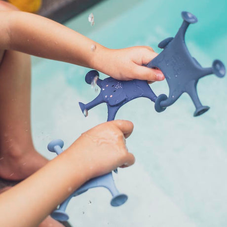 Child's hands hold blue toy starfish with suction cups above a pool. Water droplets splash, evoking a playful and cheerful atmosphere.