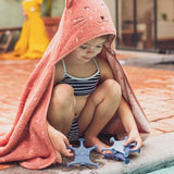 A young child in a striped swimsuit crouches by a pool, wrapped in a cozy pink towel with a cat face. They play with blue star-shaped toys.