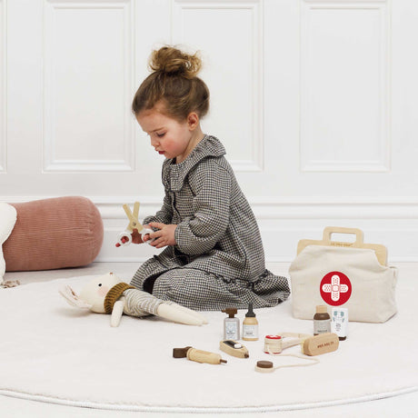 Young girl using toy wooden scissors on a plush mouse next to a vet play kit on a white rug.