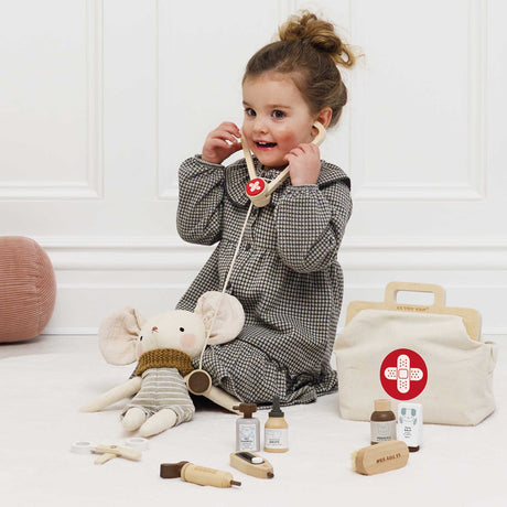 Young girl playing vet with a wooden stethoscope on a plush mouse toy next to a medical kit.