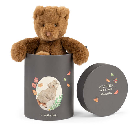 Brown teddy bear sitting in a cylindrical grey gift box by Moulin Roty, with the lid propped up next to it.