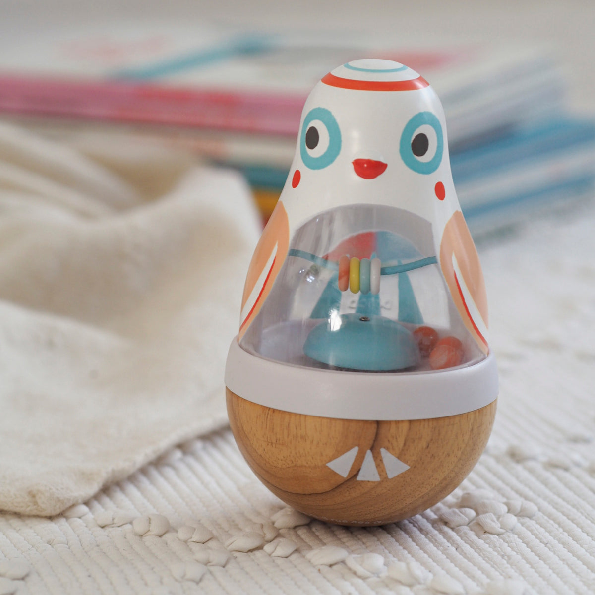 Colorful owl roly-poly wobbler baby toy with wooden base and internal rattle beads on a white textured rug.