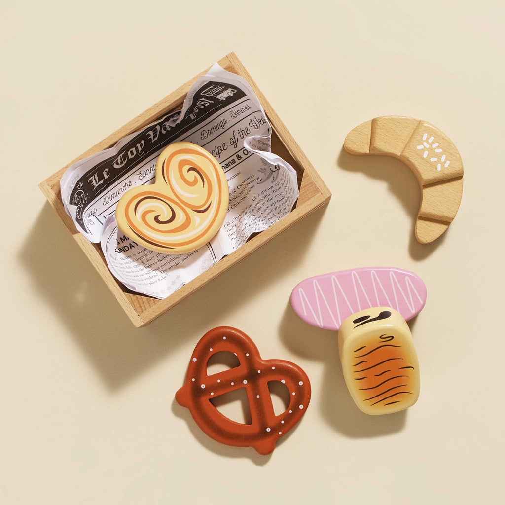 A wooden toy crate labeled "Le Toy Van" contains colorful, wooden bakery items, including a pretzel, croissant, and heart-shaped cookies, evoking a playful and cheerful mood.