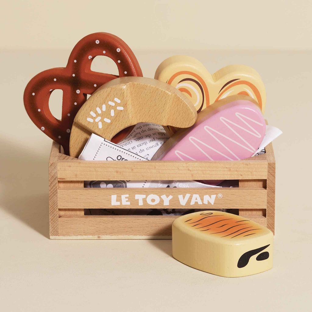 A wooden toy crate labeled "Le Toy Van" contains colorful, wooden bakery items, including a pretzel, croissant, and heart-shaped cookies, evoking a playful and cheerful mood.