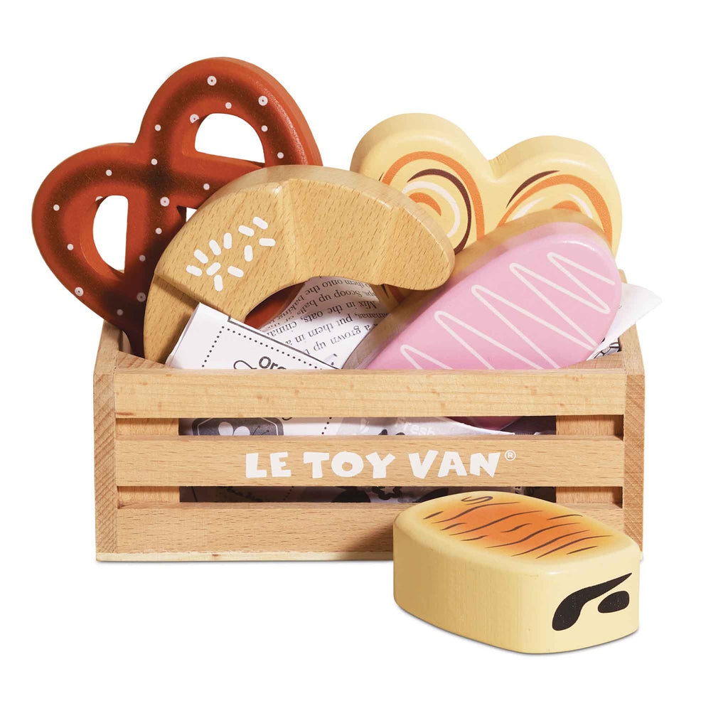 A wooden toy crate labeled "Le Toy Van" contains colorful, wooden bakery items, including a pretzel, croissant, and heart-shaped cookies, evoking a playful and cheerful mood.