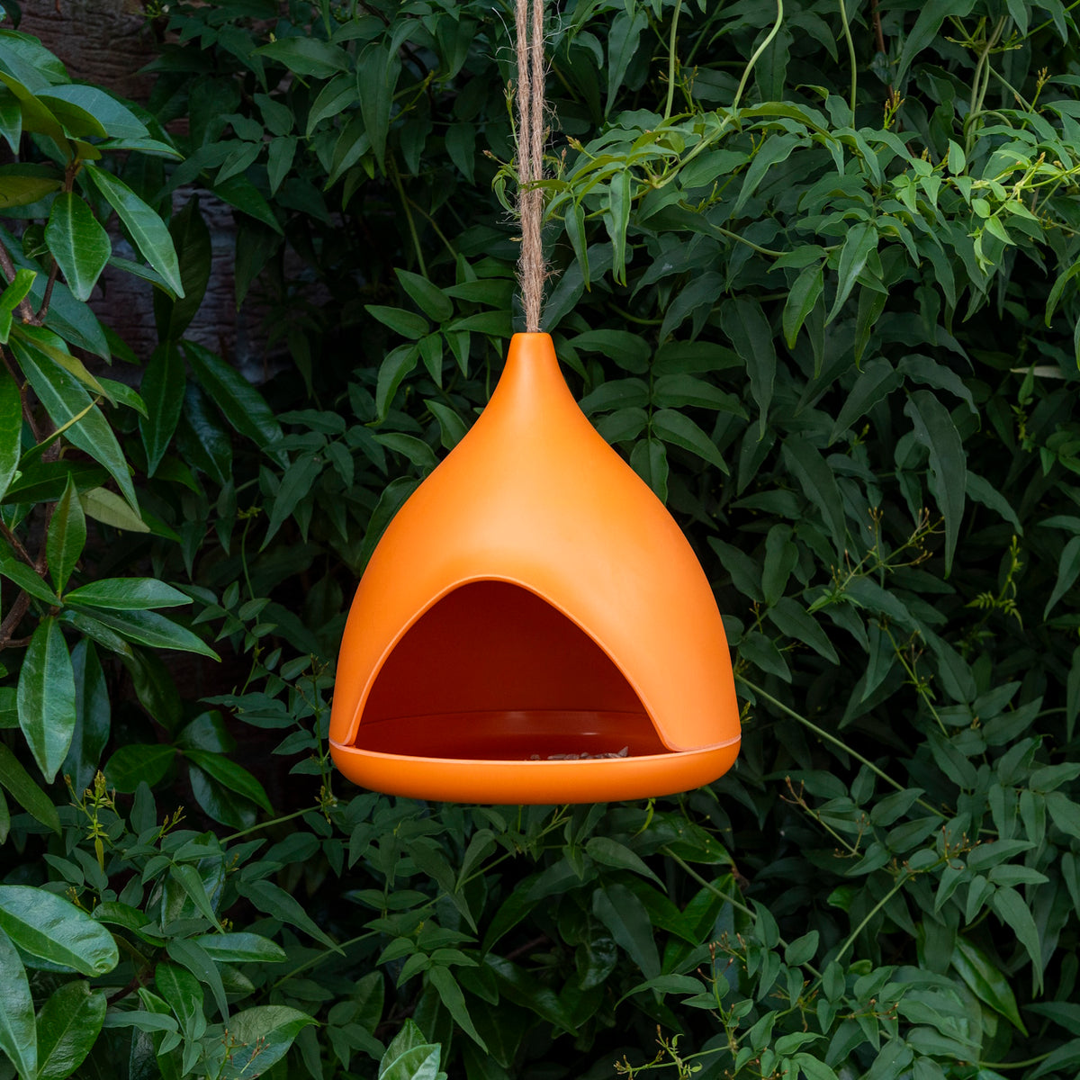 Modern orange bamboo fibre bird feeder hanging from a twine rope against a lush green leafy garden background.