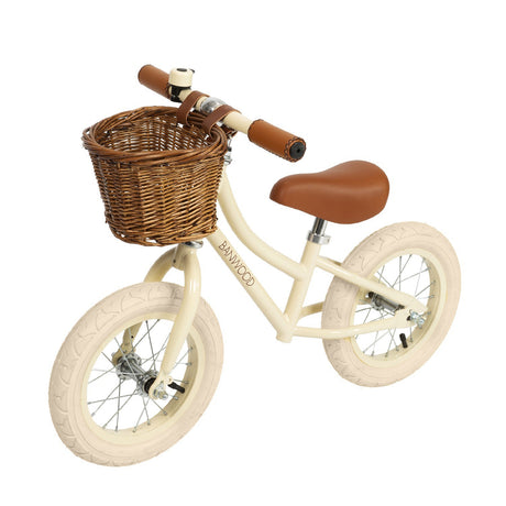 A stylish kids' balance bike with a cream-colored frame, adjustable saddle and handlebars, and a classic wicker basket for timeless charm.