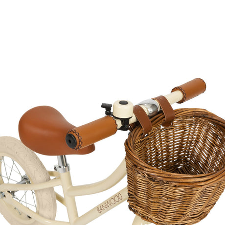 A stylish kids' balance bike with a cream-colored frame, adjustable saddle and handlebars, and a classic wicker basket for timeless charm.