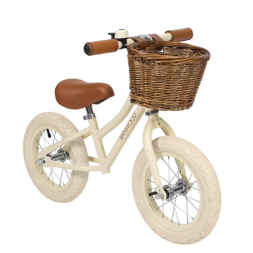 A stylish kids' balance bike with a cream-colored frame, adjustable saddle and handlebars, and a classic wicker basket for timeless charm.
