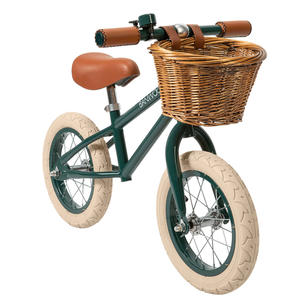 A vintage-style kids' balance bike featuring a dark green frame, adjustable saddle and handlebars, and a wicker basket for added charm.