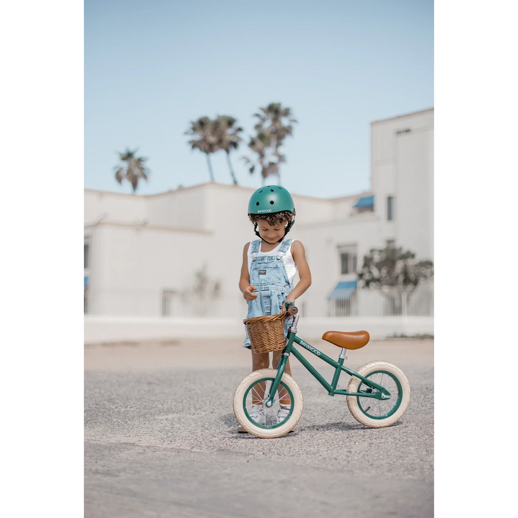 A vintage-style kids' balance bike featuring a dark green frame, adjustable saddle and handlebars, and a wicker basket for added charm.