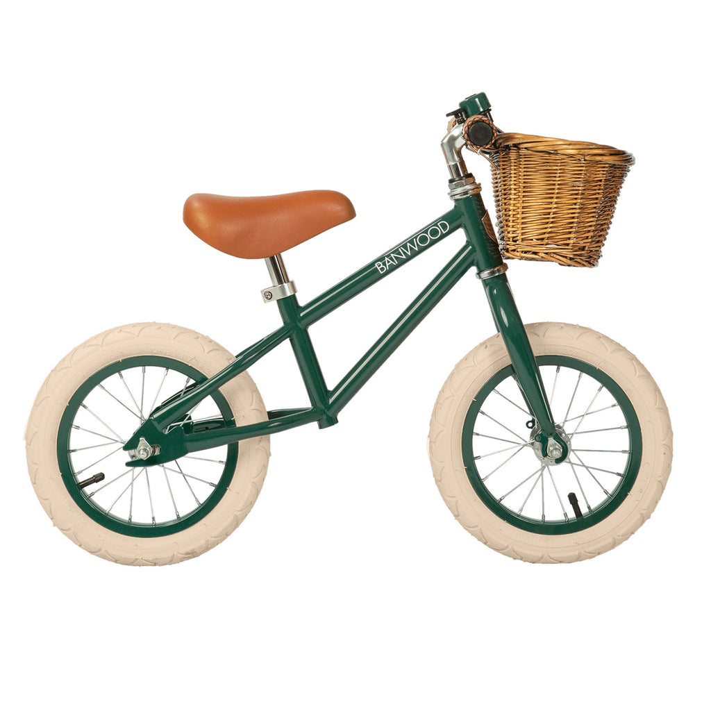 A vintage-style kids' balance bike featuring a dark green frame, adjustable saddle and handlebars, and a wicker basket for added charm.