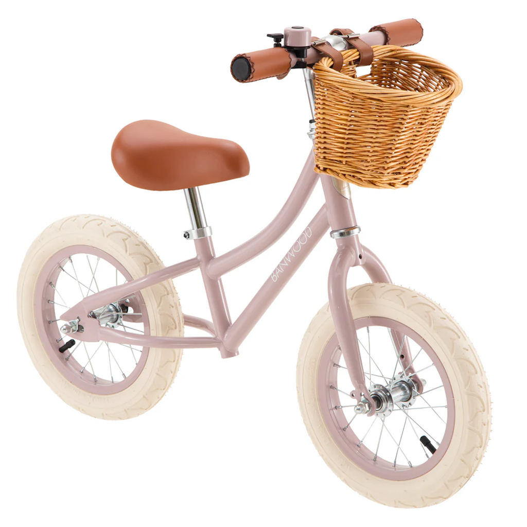 Pink balance bike with cream tires, a brown seat, and handlebars. It features a wicker basket on the front, conveying a playful and vintage feel.