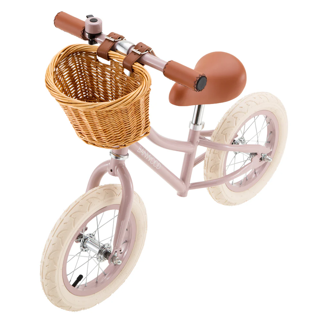 Pink balance bike with brown leather seat and grips, featuring a wicker basket on the front. The bike has white tires and a minimalist design.