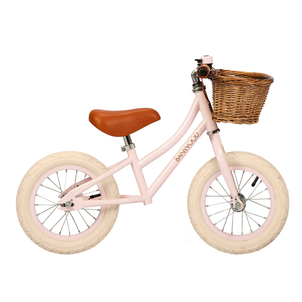 A charming vintage-inspired balance bike for kids, featuring a pastel pink frame, adjustable seat and handlebars, and a classic wicker basket.