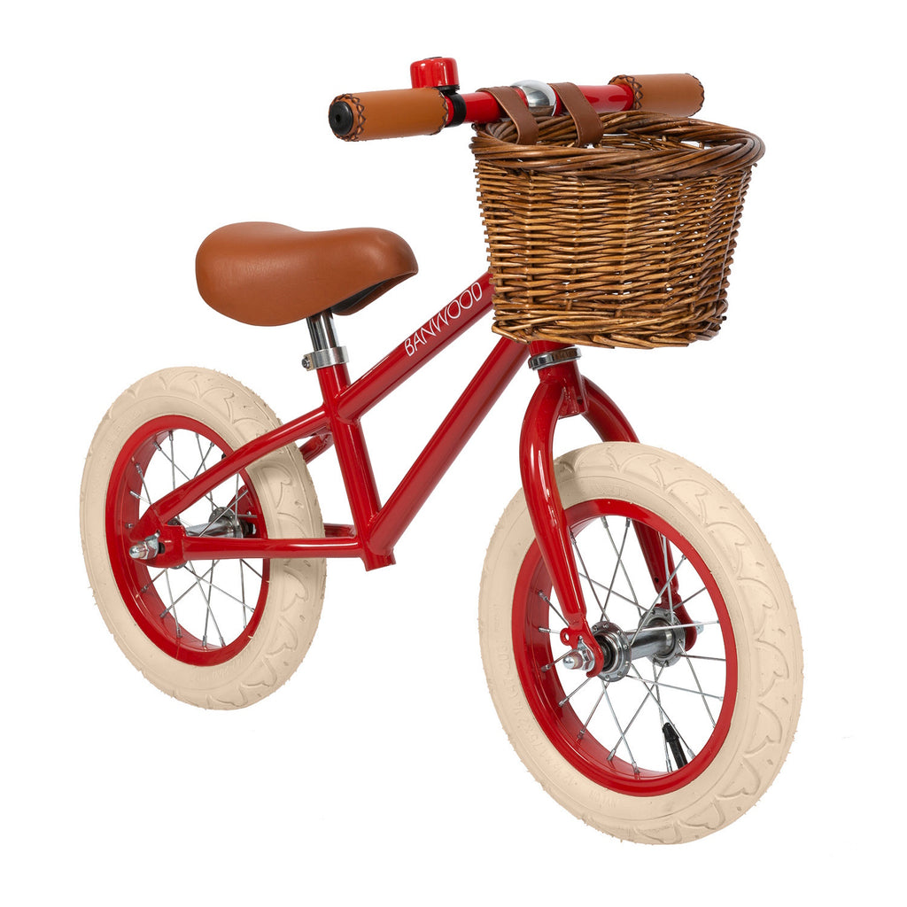 A vintage-style children’s balance bike featuring a bold red frame, adjustable seat and handlebars, and a timeless wicker basket.
